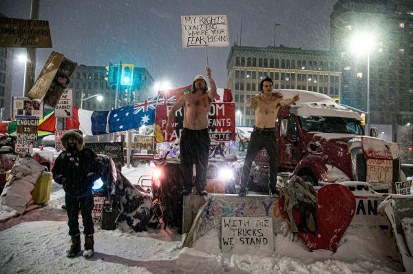 Two shirtless young men dance and sing on a concrete divider in front of a second concentration of vehicles and protesters at the top of Rideau Street, approximately 500 meters from the Canadian Parliament Buildings.